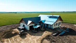 House under construction in a wide rural field, with a metal roof being installed and a worker in an orange shirt on the roof; construction equipment and materials nearby.