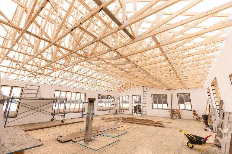 Interior of a building under construction with exposed wooden roof trusses and scaffolding visible through windows