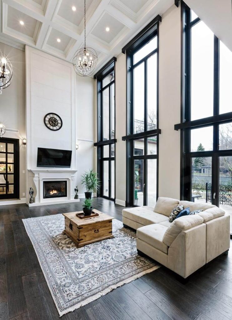 Spacious living room with a coffered ceiling, tall black-framed windows, a white fireplace, beige sectional, and a wooden coffee table.